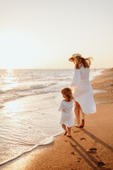 A little girl and a young mother in white dresses and a hat walk barefoot on the sand on the ocean...