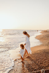 A small child, a girl in her mother's arms, both in white dresses, walk barefoot along the ocean at sunset.