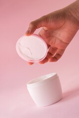 Vertical shot of a hand opening a container of skin cream on a pink background