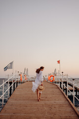 A young girl with long hair in a white dress walks towards the sea along a wooden pier and holds a hat in her hands. Hair develops the wind