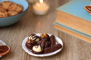 Books, reading glasses, e-reader, plate of chocolate pralines, bowl of cookies, cups of tea and lit candles on the table. Selective focus.