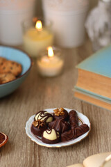 Books, reading glasses, e-reader, plate of chocolate pralines, bowl of cookies, cups of tea and lit candles on the table. Selective focus.