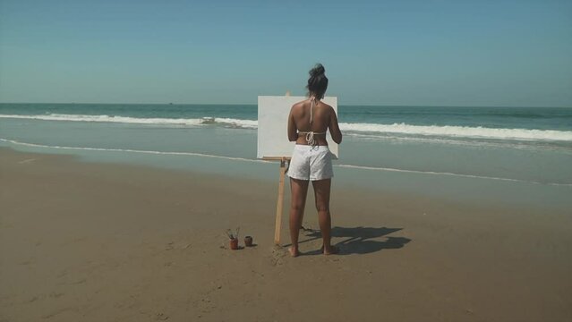 Cinematic backshot, a young female is painting a beautiful canvas at a sunny beach.