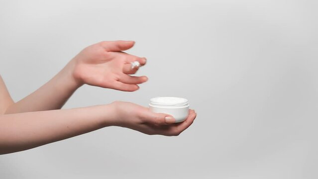 Woman is holding jar of face cream in her hand, taking a sample with a finger and applying it on a hand, gray background, copy space on the right. 4k, slow motion.