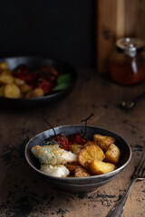 Roasted potatoes and tomatoes with codfish in a black plate. Rustic food