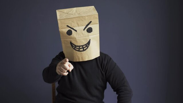 A man in a black turtleneck with a bag on his head, with an angry smiley face drawn, emotionally gloats while sitting on a chair. Emotions and gestures.