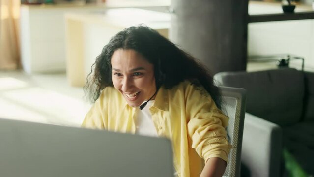 Excited emotional young hispanic woman gamer sitting at desk and playing in video games on computer at home Happy player female take part in online competition and talking to players online indoors