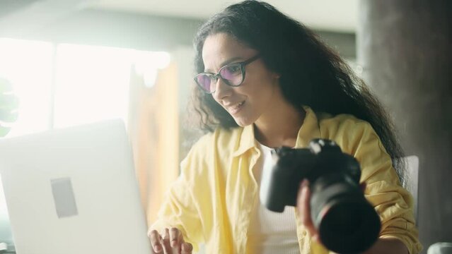 Portrait of pretty young hispanic woman photographer hold digital camera looking at screen choosing photos for editing while sitting in front of laptop computer at home workplace