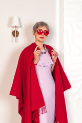 A fashionable elderly lady in a pink dress and a large scarf is draped over her shoulders is posiang against a white background.