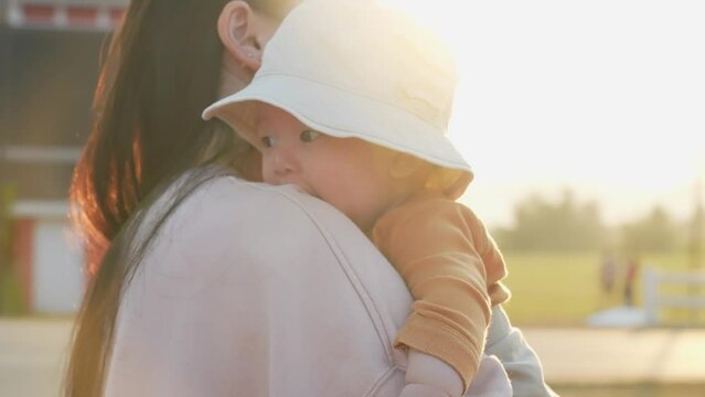 Asian mother carry up her infant baby child during sunset golden hour at outdoor recreation parkland, warmness love from mommy, family new generation member, hug and take care of newborn son