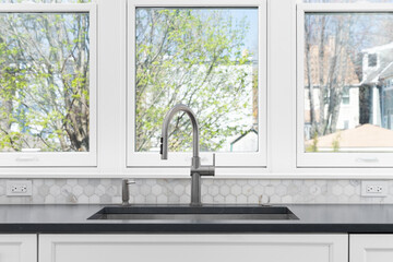 A kitchen sink detail with white cabinets, grey countertop, a marble hexagon backsplash, and large,...