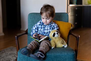 Adorable fair toddler boy sitting in armchair looking down while turning the pages of his book