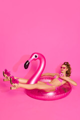 Portrait of blond girl wearing swimsuit holding donut and laying on inflatable flamingo over pink studio background. Pool party. Summer time cheeling