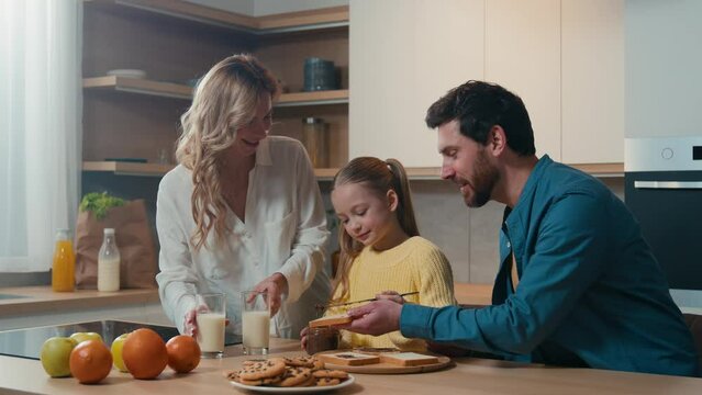 Happy Caucasian family preparing breakfast together talking at home kitchen mother hold glasses with milk little daughter child girl prepare toasts bread with chocolate pasta with father food delivery