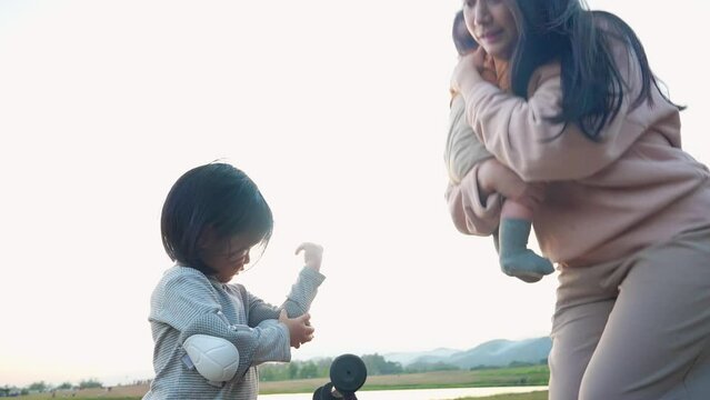 Caring asian young mother takes care of her children at natural park. Caring mommy holding infant helping little adorable daughter wearing elbow pads. Family Picnic in Park. Active Activity. Love Bond
