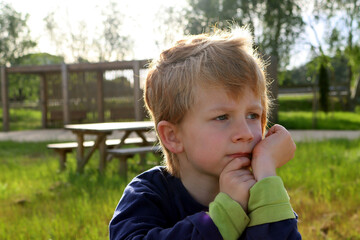 A blond boy with sad eyes looks into the distance. A pensive five-year-old boy in the rays of the sun.