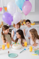 cheerful kids with balloons looking at birthday girl near cake and party horns on table.