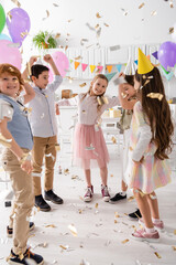 happy kids in party caps dancing under falling confetti during birthday celebration at home. - obrazy, fototapety, plakaty