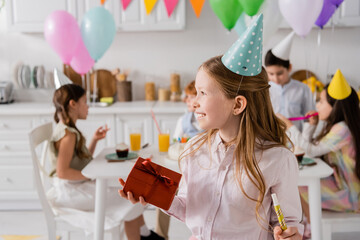 happy preteen girl holding birthday gift box and party horn near friends on blurred background.