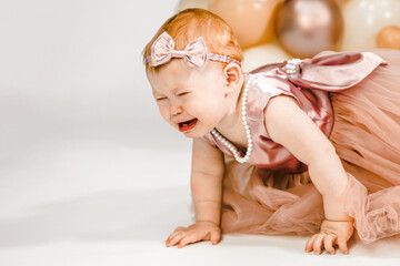 Little crying unhappy redhead baby girl celebrates first birthday anniversary. 1 year family party Professional photoshoot in photo studio. Cute adorable red hair kid in pink dress.Home children room