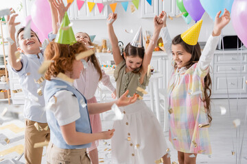 group of joyful kids in party caps dancing under falling confetti during birthday celebration at...