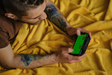 Guy Uses Green Mock-up Screen Smartphone. Young man lies inside on yellow blanket on bed and plays using phone and joystick, top view. Hands with multicolored tattoos close-up.