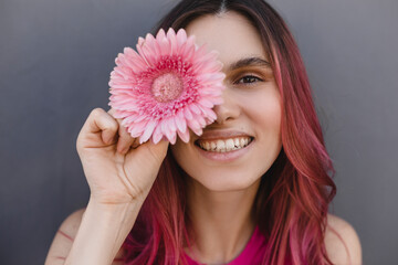 Close up shot of cheerful smiling attractive young pink hair woman covering a part of her face with pink gerbera flower, isolated over grey background. Women cosmetics and face skin care. Happy girl