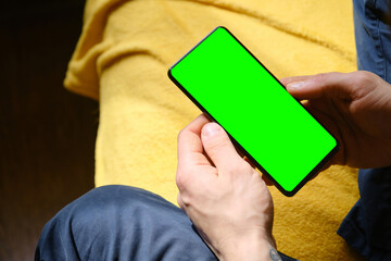 Young man sitting inside on yellow blanket on bed and uses phone to work or browse the Internet online, top view. Hands close-up. Guy Uses Green Mock-up Screen Smartphone.