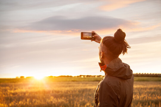 a boy from behind with long hair with a bow making photos with the mobile in the field looking at the sun in the sunset with the golden sky and with clouds