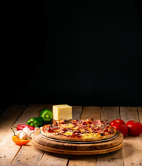 Pizza surrounded by peppers, tomatoes, mushrooms and a block of mozzarella cheese on a rustic wooden table against a black background.