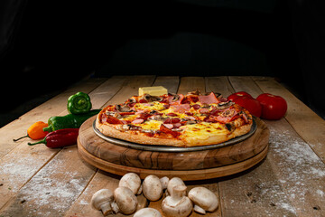 Pizza surrounded by peppers, tomatoes, mushrooms and a block of mozzarella cheese on a rustic wooden table against a black background.