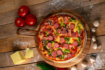Pizza surrounded by peppers, tomatoes, mushrooms and a block of mozzarella cheese on a rustic wooden table against a black background.