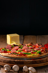 Pizza surrounded by peppers, tomatoes, mushrooms and a block of mozzarella cheese on a rustic wooden table against a black background.