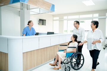 Asian family with grandmother, husband and pregnancy wife on wheelchair move pass area near reception or counter in hospital and greeting by Caucasian nurse.