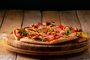 Pizza surrounded by peppers, tomatoes, mushrooms and a block of mozzarella cheese on a rustic wooden table against a black background