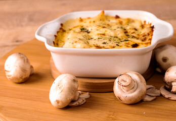 Baked lasagna or pasta gratin in a white ceramic tray surrounded by mushrooms on a rustic wooden table on a black background