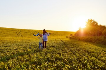 Fototapeta premium Running boy and girl holding two yellow and blue airplanes toy in the field during summer sunset. Kids dreams of flying and aviation.