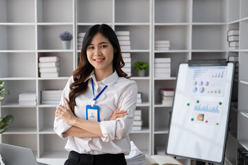 Portrait of young smiling Asian businesswoman looking at camera with crossed arms, standing in the office room.