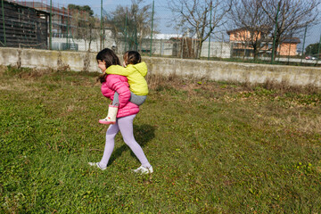 younger child do piggyback ride on elder sister outdoors in bright jackets