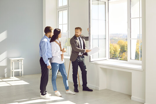 Handsome bearded male realtor in suit with documents in his hands advises young couple before buying new house, demonstrates view from the window. Three people are standing in modern sunny apartment.