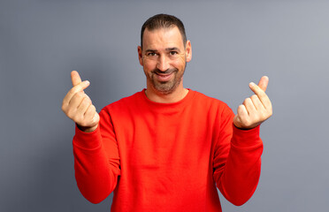 Bearded hispanic man in his 40s wearing a red sweater making the money gesture rubbing his fingers with a greedy face, isolated over gray background.