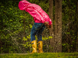 Child in bright pink parka and tan rain boots, looking down and splashing in rain puddle