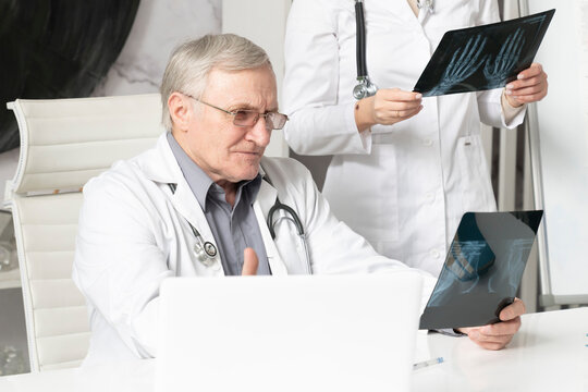 Portrait of aged doctor sitting at the desk and holding x-ray image and his young female colleague standing close to him