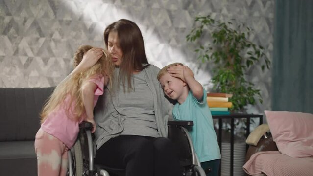 Happy mother with paralysis sitting in wheelchair hugs and kisses little son and daughter. Woman expresses love to children spending time at home
