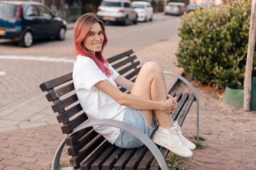 Fototapeta na wymiar Woman with pink hair sitting on bench and smiling, walking in park, looking carefree and happy. Modern girl breathing fresh air on a walk, look free and joyful.
