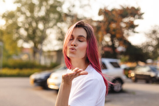 Pensive pink hair woman turn side and make blow kisses walking on the street. Outdoor shot of happy hippie lady in boho freedom style. Girl send air kiss to camera, closed eyes, love concept.