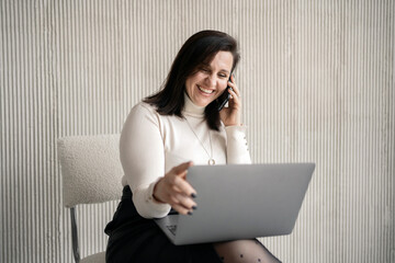 A woman using a laptop and a phone happily smiles and shows her teeth.