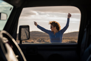 Excited happy female traveler jumping crazy having fun at arrival destination viewed from inside the car. Driver pov of woman dancing outside happy. Freedom and joy lifestyle people loving travel