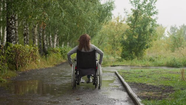 Woman with paralysis rests in city park moving along asphalt path with puddles on windy spring day. Restoring health in nature backside view