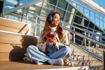 Optimistic woman wearing headphones and holding phone outdoors. Young woman listens to music, audiobook or podcast while walking on sunny street. Communication in social networks. Lifestyle.